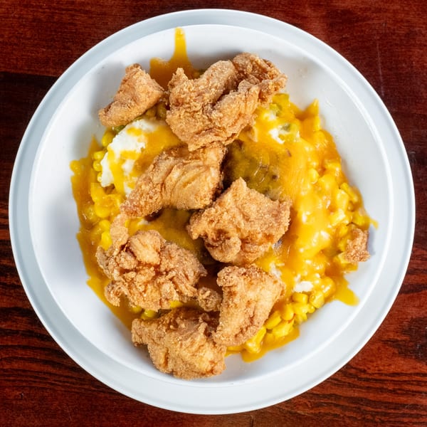 Fried chicken pieces over mashed potatoes and corn with yellow sauce in a white bowl.