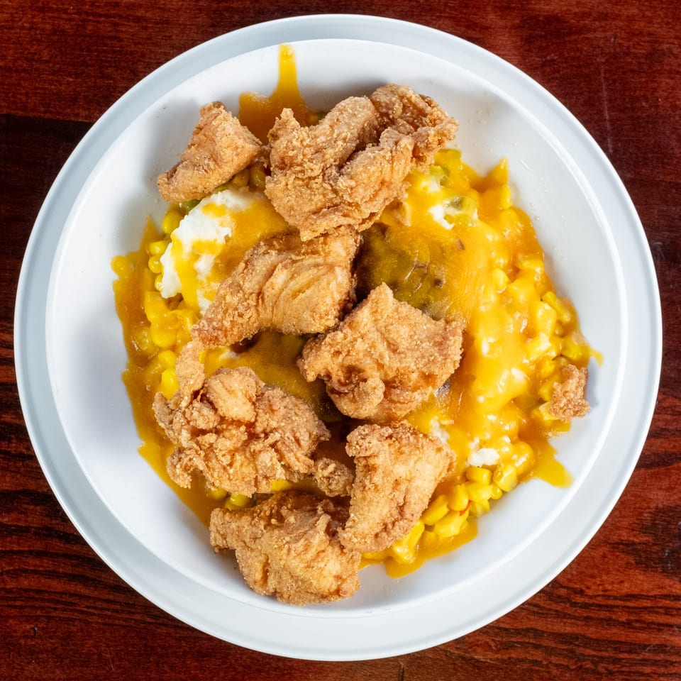Fried chicken pieces over mashed potatoes and corn with yellow sauce in a white bowl.