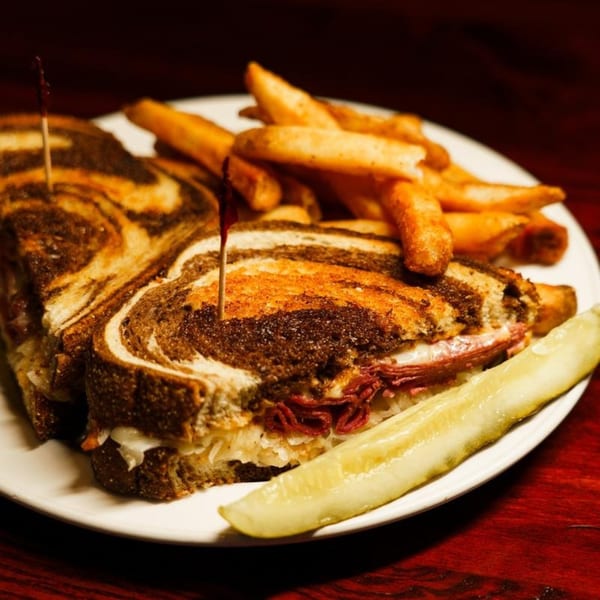 Bullwinkle's Top Hat Bistro's Reuben sandwich with fries and a pickle spear on a white plate.