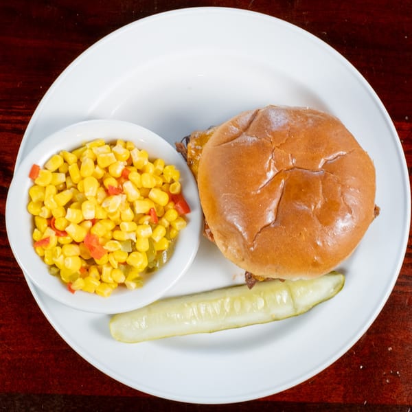 BBQ sandwich on a bun with a side of corn and red peppers, and a pickle spear.