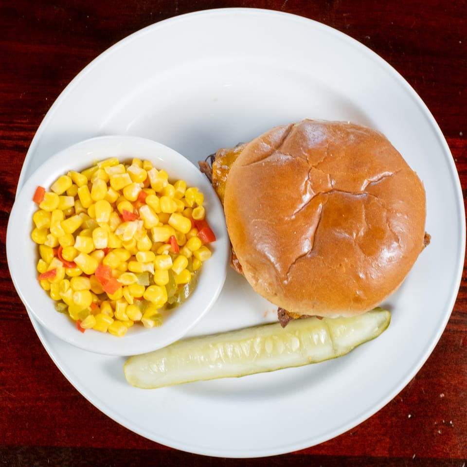 BBQ sandwich on a bun with a side of corn and red peppers, and a pickle spear.