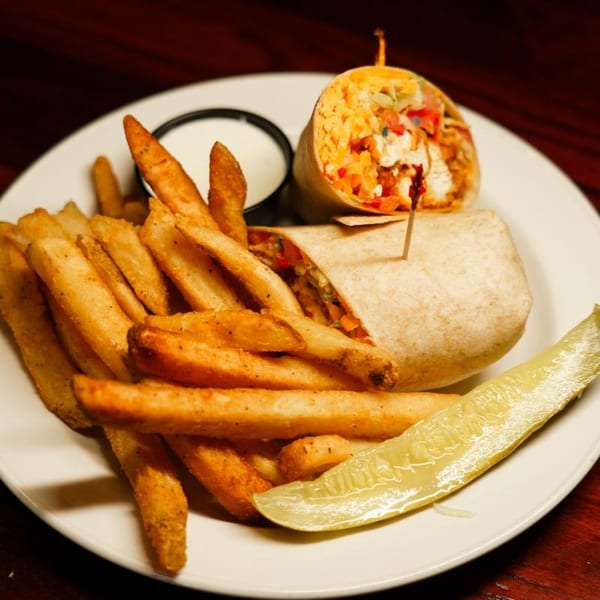 A BBQ chicken wrap, golden fries, a side of ranch, and a pickle spear on a white plate.