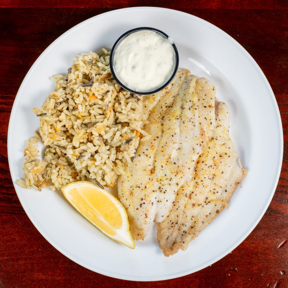 Grilled fish fillet with rice pilaf, a lemon wedge, and tartar sauce on a white plate.
